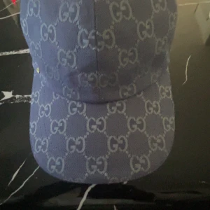 Dubai blue gg keps med gg mönster  - Snygg blå keps från Gucci med det klassiska GG-mönstret över hela kepsen. Kepsen har en böjd skärm och ett litet guldfärgat GG-emblem på sidan. Materialet är robust och känns lyxigt, insidan är ljus och mjuk mot huvudet. Perfekt för dig som gillar designer och vill sticka ut.