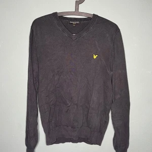 Mörkgrå Lyle & Scott tröja v-ringad - Snygg mörkgrå v-ringad tröja från Lyle & Scott med deras klassiska gula fågel-logga broderad på bröstet. Tröjan är långärmad och har ribbade muddar vid ärmslut och nederkant. Perfekt för lager-på-lager och stilren look.