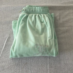 Mintgröna mjukisbyxor Juicy Couture - Supermjuka mintgröna mjukisbyxor från Juicy Couture med glittrande JC-logga i strass på ena benet. Byxorna har snörning i midjan, raka ben och är tillverkade i ett skönt velourmaterial. Perfekta för en chill och trendig look.