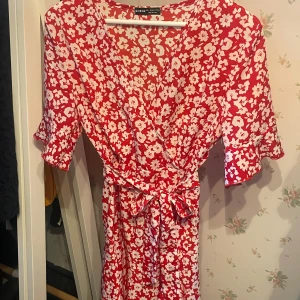 Röd blommig omlottklänning från SHEIN - Superfin röd kortklänning från SHEIN med vita blommor, omlottmodell och knytband i midjan. Klänningen har v-ringning, volangdetaljer på ärmarna och är kortärmad. Perfekt för sommarens alla tillfällen!