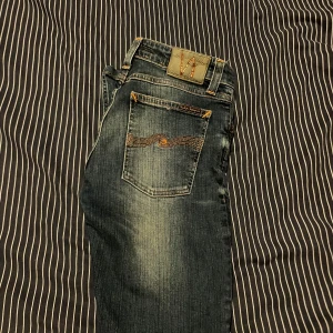 Blå jeans från Nudie Jeans, W28 L32 - Säljer ett par klassiska blå jeans från Nudie Jeans med snygga slitningar och kontrastsömmar. Modellen har raka ben och femficksdesign med Nudie-logga på bakfickan. Jeansen är i bomull och har orange detaljer vid sömmarna.