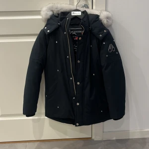 Svart parkas Moose Knuckles med päls - Snygg svart parkas från Moose Knuckles med lyxig grå päls runt huvan. Jackan har silverfärgade detaljer, dragkedja och knappar framtill samt logga på ärmen. Insidan har ett broderat motiv med hockeyspelare. Perfekt för kalla dagar. Köpt på Nk barnavdelning, använd fåtal gånger ungefär 3-4. OBS Storlek är i barnstorlek inte here 