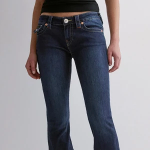 Mörkblå bootcut jeans från True Religion - Slutsålda överallt!!! Snygga mörkblå jeans från True Religion med bootcut passform och låg midja. Jeansen har klassiska kontrastsömmar, dekorativa bakfickor med lock och knappar samt en tydlig logopatch bak. Perfekta för dig som gillar en tidlös och cool look.