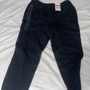 Svarta Nike mjukisbyxor med snörning - Svarta mjukisbyxor från Nike med dragsko i midjan och diskret Nike-logga på vänster ben. Byxorna har dragkedjeförsedda fickor och är tillverkade i mjukt bomullsmaterial. Perfekta för chill eller träning, med en slim passform och sportig look.