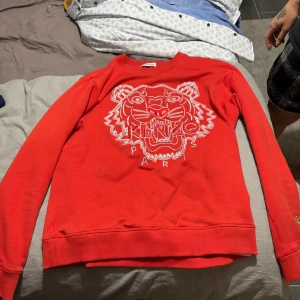 Röd Kenzo sweatshirt med tigertryck - Säljer en röd sweatshirt från Kenzo med ikoniskt broderat tigerhuvud och Kenzo Paris-text på bröstet. Tröjan har rund hals, långa ärmar och ribbade muddar. Perfekt statement-piece för dig som gillar streetwear och snygga detaljer.