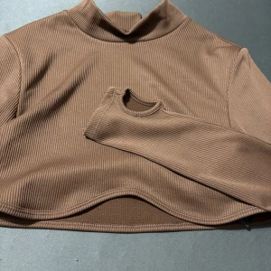 Brun ribbad croppad polotopp - Säljer en brun ribbad croppad polotopp med lång ärm. Toppen har en hög hals och är tillverkad i ett stretchigt material som sitter snyggt på kroppen. Perfekt att styla med jeans eller kjol för en trendig look.