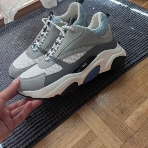 Chunky sneakers från Dior i grått och vitt - Säljer ett par chunky Dior sneakers i grått, vitt och blått. Skorna har meshdetaljer, skinnpaneler och en kraftig vit sula med svarta och blå inslag. Snygg snörning och sportig vibe, perfekt för dig som gillar statement sneakers.