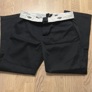 Svarta Dickies byxor med vit midja - Svarta byxor från Dickies med en unik vit midja och svart spänne. Klassisk rak passform med sidfickor och bakficka med knapp. Perfekta för dig som gillar streetwear och vill ha något som sticker ut lite extra.