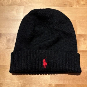 Svart stickad mössa från Polo Ralph Lauren - Svart stickad mössa från Polo Ralph Lauren med klassisk röd broderad logga framtill. Mössan har uppvikt kant och är tillverkad i mjukt material som håller dig varm under kyliga dagar. Perfekt accessoar för en stilren look.