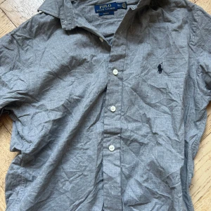 Grå skjorta från Polo Ralph Lauren - Snygg grå skjorta från Polo Ralph Lauren i slim fit-modell. Skjortan har klassisk krage, knäppning framtill med vita knappar och den ikoniska broderade loggan på bröstet. Perfekt för dig som gillar stilrena och tidlösa plagg.
