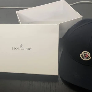 Svart Moncler keps med broderad logga - Snygg svart keps från Moncler med klassisk böjd skärm och broderad Moncler-logga framtill i vitt och rött. Kepsen har en stilren design och är tillverkad i bomull för en bekväm känsla. Perfekt för dig som vill ha en exklusiv och trendig accessoar.