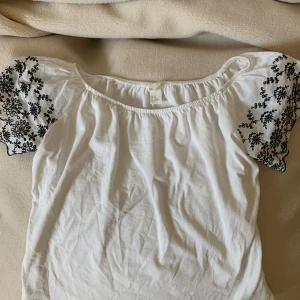 Vit blus med broderade puffärmar H&M XS - Superfin vit blus från H&M i storlek XS med puffiga kortärmar som har svart broderat blommönster. Blusen har en vid, rundad halsringning och är gjord i mjuk bomull.