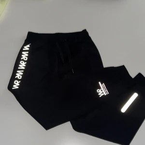 Svarta mjukisbyxor från Lindex Kids - Svarta mjukisbyxor från Lindex Kids med vita WR-tryck längs benet och reflexdetaljer. Byxorna har resår i midjan med snörning och är tillverkade i mjukt material, perfekta för chill eller träning.