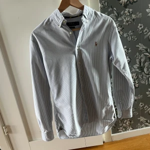 Randig skjorta från Polo Ralph Lauren - Säljer en slim fit skjorta från Polo Ralph Lauren i vitt och blått med tunna ränder. Skjortan har klassisk krage, knappar framtill och lång ärm. Perfekt för dig som gillar stilrena och tidlösa plagg.