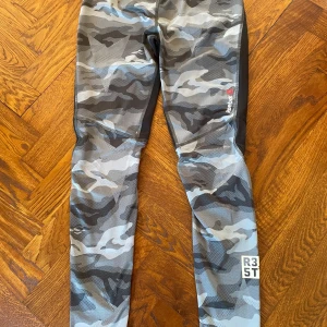 Reebok camo tights med ACTIVCHILL - Snygga träningstights från Reebok med ACTIVCHILL-teknologi. Byxorna har grått och svart camouflagemönster, svarta paneler på baksidan och tryckdetaljer vid benet. Perfekta för dig som vill sticka ut på gymmet eller löprundan.