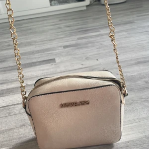 Beige axelväska från Michael Kors - Snygg beige axelväska från Michael Kors med guldfärgad kedja och justerbar axelrem. Väskan är i skinnimitation och har dragkedja upptill samt en liten ficka med dragkedja på baksidan. Perfekt storlek för det viktigaste och stilren design. PRIS KAN DISKUTERAS!!