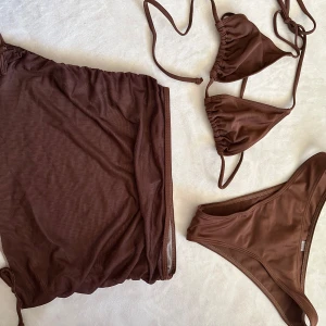 Brun bikini med kjol och knyt - Supersnygg brun bikini med trekantsöverdel och knyt i nacken samt matchande bikinitrosa. Setet kommer med en meshkjol i samma färg som ger extra stilpoäng på stranden. Perfekt för dig som vill ha en trendig och enkel look i sommar. Oanvänd men tvättad i tvättmaskin!