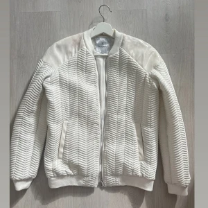 Vit jacka från Kookai - Säljer en riktigt snygg vit bomberjacka från Kookai. Jackan har mönster, glansiga detaljer på axlarna och dragkedja fram! Jackan har någon färgskiftning på ena axeln men inget markant