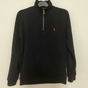 Svart half zip tröja från Polo Ralph Lauren - Svart långärmad tröja från Polo Ralph Lauren med röd broderad logga på bröstet. Tröjan har en hög krage med half zip-dragkedja och är tillverkad i mjuk bomull. Perfekt för en clean och stilren look.