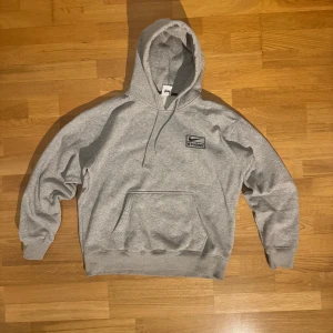 Grå Nike x Stussy hoodie i bomull - Säljer en grå hoodie från Nike x Stussy med broderad logga på bröstet. Tröjan har huva med snörning, känguruficka och ribbade muddar. Tillverkad i mjuk bomull, perfekt för chill dagar. Klassisk och clean streetwear-look.