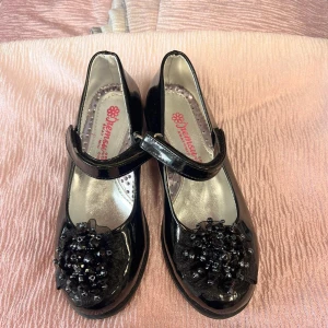 Svarta lack ballerina med pärldetalj - Snygga svarta ballerina med glansig lackyta och rund tå. Rem över foten med kardborre för enkel påtagning. Dekorerade med en stor svart rosett och pärlor framtill som ger extra bling. Perfekt för dig som gillar att sticka ut med detaljer.