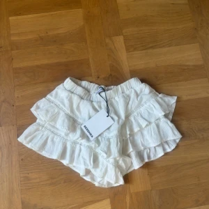 Vita volangshorts från Bershka - Säljer ett par vita shorts från Bershka med flera lager volanger och elastisk midja. De har en luftig och flowy känsla, perfekta för varma dagar. Materialet är lätt och känns som bomull. Supersöta och trendiga för sommaren!