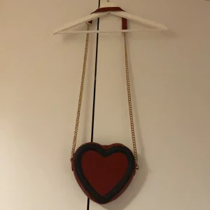 Hjärtformad röd axelväska med kedja - Superfin hjärtformad axelväska i rött med mörk kant och guldfärgad kedjerem. Perfekt accessoar för att sticka ut och ge din outfit en söt touch. Väskan har unik form och är lätt att bära över axeln. Lite trasig 