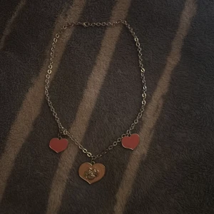 Halsband med röda hjärtan och motiv - Kedjehalsband i metall med tre hjärtformade hängen, två i röd emalj och ett större i guld med ett ingraverat motiv av en tecknad figur. Perfekt accessoar för dig som gillar söta detaljer och unika smycken.