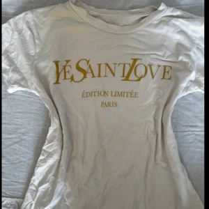Vit t-shirt Yesaintlove Paris - Vit t-shirt från Yesaintlove med guldfärgat tryck 'Édition Limitée Paris' på bröstet. Klassisk rund halsringning och korta ärmar. Tillverkad i 100% bomull, skön och lätt att bära. Perfekt statement-plagg för dig som gillar unika prints.