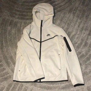 Vit Nike vindjacka med huva - Snygg vit vindjacka från Nike med svarta detaljer och dragkedja hela vägen. Jackan har huva, svarta linjer över bröstet och ärmen samt en ficka med dragkedja på ärmen. Perfekt för dig som gillar sportig och clean stil.