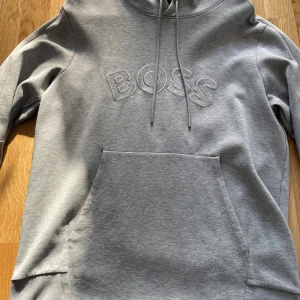 Grå hoodie från BOSS med broderad logga - Snygg grå dress från BOSS med broderad logga på bröstet. Dressen har huva med dragsko, stor magficka och ribbade muddar. Tillverkad i mjuk bomullsblandning, perfekt för chill eller streetstyle. Enkel och stilren design med en modern vibe.