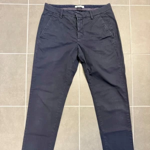 Mörkblå chinos från Dondup, stl 32 - Snygga mörkblå chinos från Dondup i klassisk rak modell. Byxorna har sidfickor, bälteshällor och bakficka med knapp och liten metall-logga. Tillverkade i bomull för en skön och stilren känsla. Perfekta för en clean och avslappnad look.