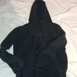 Svart Niketech med dragkedja - Mysig svart hoodie i fleece med hel dragkedja framtill och huva. Perfekt för kyliga dagar och enkel att matcha med andra plagg. Klassisk modell med långa ärmar och mjukt material.