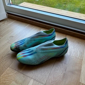 Adidas blå fotbollsskor med regnbågsdetaljer - Säljer ett par lätta fotbollsskor från Adidas i ljusblått med regnbågsfärgade detaljer och klassiska tre ränder på sidan. Skorna har en strömlinjeformad design, plös utan snörning och mönstrad ovandel. Perfekta för dig som vill sticka ut på planen.