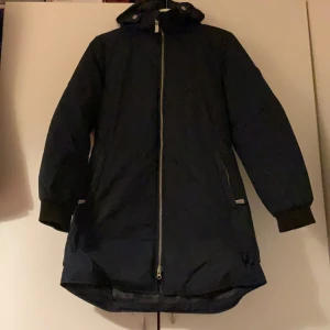 Svart lång parkajacka med huva - Snygg mörkblå parkajacka med huva och långa ärmar. Jackan har dragkedja framtill, ribbade muddar vid ärmslut och en längre passform som täcker rumpan. Perfekt för kalla dagar när du vill hålla dig varm och stilren.