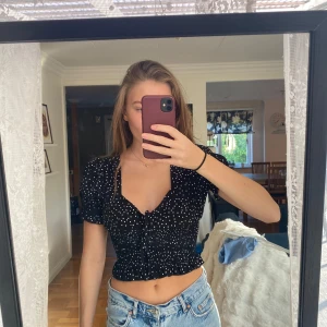 Prickig topp - Supersöt svart blus med vita prickar, croppad modell och smockad midja. Korta puffärmar och v-ringning med knyt framtill. Storlek S i super bra skick, aldrig använd och köpt på plick står inte vart den är ifrån 