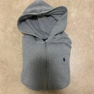 Grå hoodie från Polo Ralph Lauren - Grå hoodie från Polo Ralph Lauren med dragkedja framtill och klassisk blå logga på bröstet. Tröjan har huva, känguruficka och är tillverkad i mjuk bomullsmix. Perfekt för chill dagar eller när du vill ha en clean och enkel look.