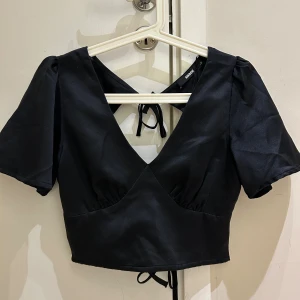 Svart omlottopp från BikBok XS - Snygg svart omlottopp från BikBok i croppad modell med korta ärmar och v-ringning. Toppen har knytband både i ryggen och i midjan för en justerbar passform. Perfekt för dig som gillar stilrena och trendiga plagg.