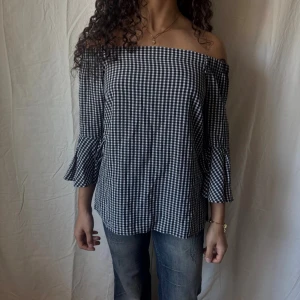 Offshoulder rutig blus med volangärm - Snygg offshoulder blus i svartvitt rutigt mönster. Blusen har trekvartsärmar med volangdetalj och rak passform. Perfekt till en trendig höstoutfit.