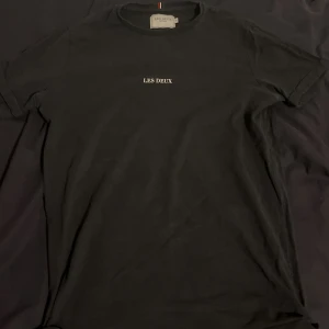 Svart t-shirt från Les Deux - Svart t-shirt från Les Deux med vit broderad logga på bröstet. Klassisk rund hals och korta ärmar. Tillverkad i mjuk bomull som känns skön mot huden. Enkel och stilren design som funkar till allt. Snygg detalj med liten röd, vit och blå rand vid nacken. Nypris ca 500-600 kr. Fråga gärna om mått. 