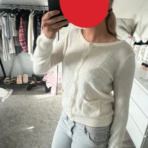 Vit kofta i ull med ficka - Mysig vit kofta i ull med rund halsringning och knappar framtill. Koftan har en liten ficka på båda  sidorna  och ribbade muddar vid ärmslut och nederkant. Perfekt att slänga på sig över en topp för en chill look.