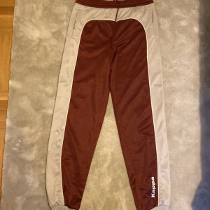 Grå och Röda mjukisbyxor från Kappa - Mjukisbyxor från Kappa med grå sidopaneler och logga på benet. Byxorna har elastisk midja med snörning. Tillverkade i mjukt syntetmaterial. Är öppen för ett lägre pris vid snabb affär. Om ni har några frågor skriv gärna!