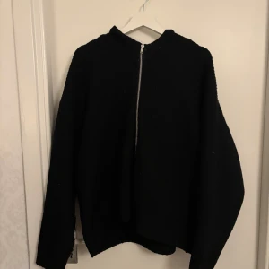 Svart stickad tröja med dragkedja - Svart stickad tröja med lång ärm och en snygg dragkedja framtill. Tröjan har en enkel, stilren design och passar perfekt till kyligare dagar. Mjuk och skön känsla, lätt att matcha med olika outfits.