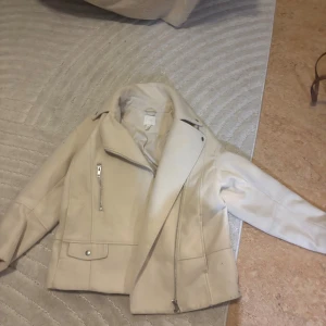 Beige skinnjacka med dragkedja - Snygg beige skinnjacka med asymmetrisk dragkedja och coola detaljer som fickor med lock och silverfärgade dragkedjor. Jackan har klassisk krage och lång ärm, perfekt för dig som vill ha en stilren look med edge.