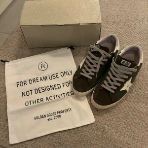 Golden Goose sneakers 44 - Snygga Golden Goose sneakers i mörkgrönt och brunt mocka med vita detaljer och ikonisk stjärna på sidan. Skorna har grå snörning, vit häl och platt sula. Perfekta för dig som gillar en cool och avslappnad stil. Vid köp inkluderar originala box och påse som tillhör skorna! Hör gärna av vid frågor! 