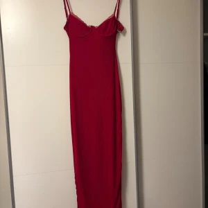 Röd långklänning med spetsdetaljer - Röd långklänning med smala axelband och spetskant vid bysten. Klänningen har figurnära passform, markerad byst och en snygg slits baktill. Materialet är mjukt och stretchigt, perfekt för en statement-look.