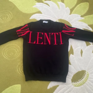 Stickad Svart Valentino tröja med röd logga - Säljer en svart tröja från Valentino med stor röd logotyp som sträcker sig över bröstet och ut på ärmarna. Tröjan har rund halsringning och är långärmad. Materialet är mjukt och stickat, perfekt för dig som gillar statement-plagg.