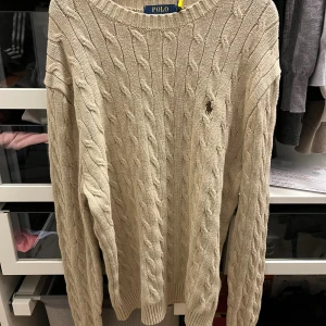 Beige kabelstickad tröja Polo Ralph Lauren - Kabelstickad beige tröja från Polo Ralph Lauren med klassisk rund halsringning och broderad logga på bröstet. Tröjan är i mjuk bomull och har långa ärmar samt ribbade muddar för en snygg passform. Helt oanvänd St S passar även M