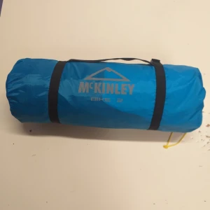 McKinley Bike 2 - McKinley Bike 2 tält i blått fodral, kompakt och lätt att bära. Perfekt för cykel- och campingturer. Fodralet ser ut att vara i gott skick utan synliga skador eller slitage. Smidig förvaring med bärhandtag och remmar.