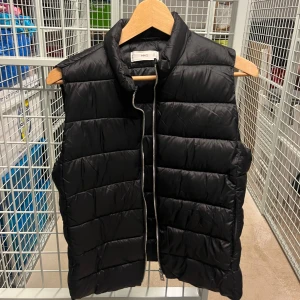 Svart dunväst från MANGO - Svart dunväst från MANGO med krage och silverfärgad dragkedja framtill. Västen har en stilren, puffad look och är tillverkad i syntetmaterial. Perfekt att slänga över en hoodie för extra värme under höst och vinter.